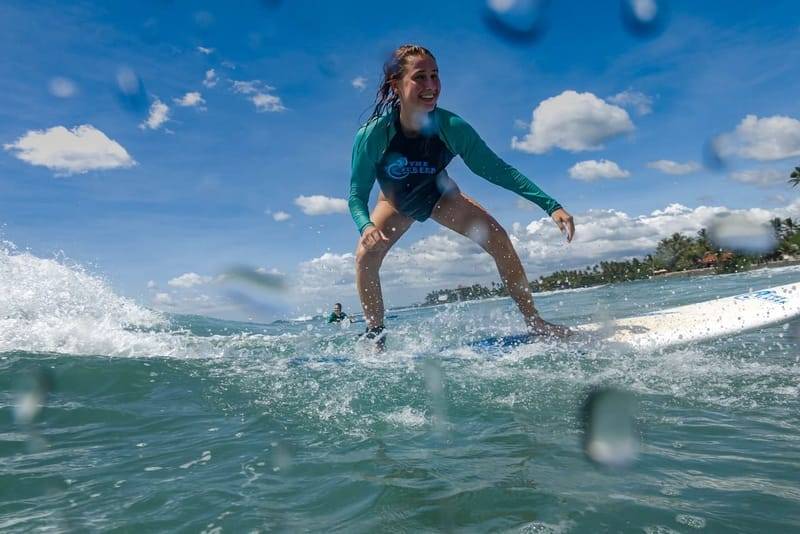A surfer standing up on a beginner wave in a teal rash guard at Weligama Bay