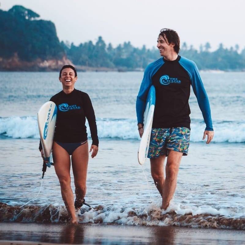 Two guests in The Surfer rash guards walking out of the sea with their boards, Weligama